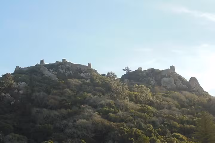 Panoramic view of the ancient Moorish Castle perched atop a lush hill under a clear blue sky in Sintra, Portugal.