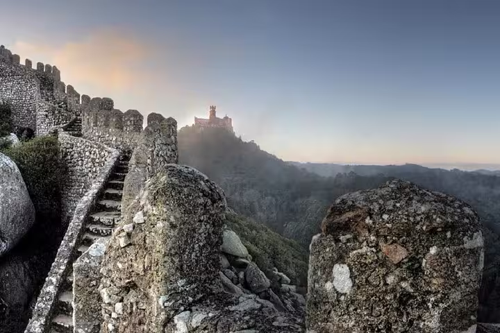 Ancient stone walls of the Moorish Castle overlook Sintra's lush landscape, with Pena Palace visible in the misty distance.