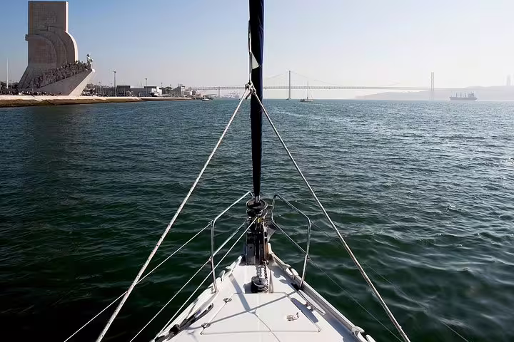 View from a sailboat's bow on the Tagus River with Lisbon's historic monuments and bridge in sight.