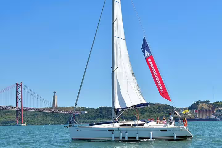Sailboat gliding on the Tagus River with Lisbon's iconic bridge and Christ the King statue in the background.