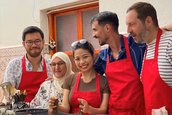 Group of five people in red aprons enjoying a cooking class with Moroccan utensils in a cozy kitchen setting.