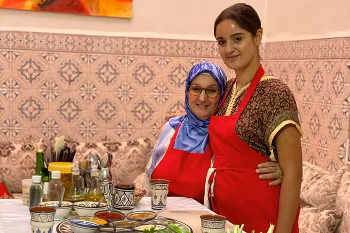 Two women in red aprons smiling beside a table set with Moroccan cooking ingredients and spices.