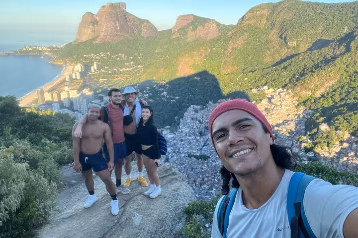 A group of hikers takes a selfie with stunning views of Vidigal and Rio's lush mountains from the Two Brothers trail.