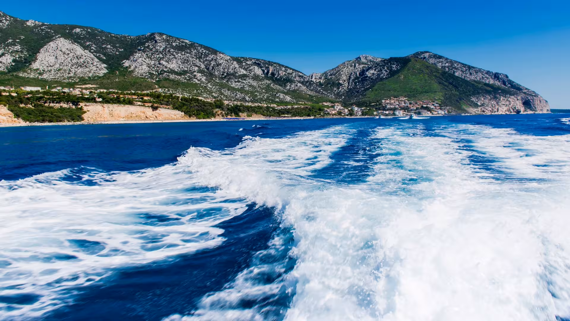 Motorboat trail on blue waters with stunning Sardinian coastline and mountains in the background, ideal for a boat tour.