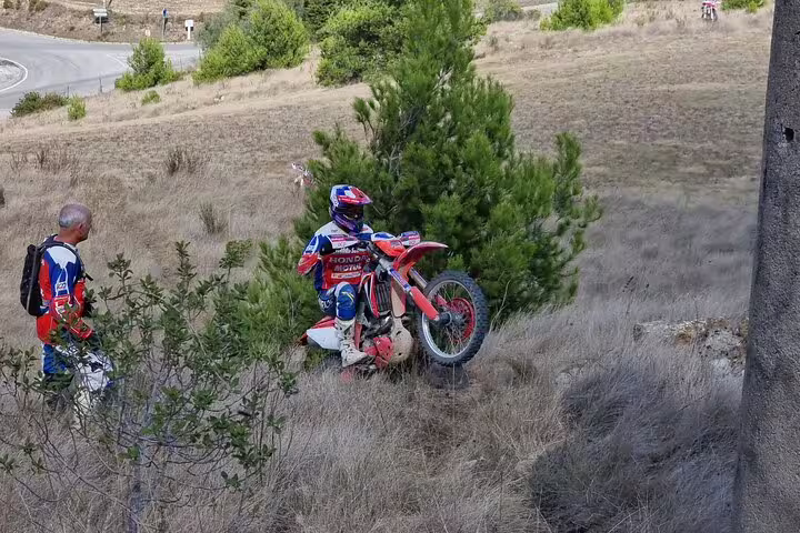 Motorcyclist performs uphill maneuver on Lisbon enduro tour, watched by fellow rider amidst greenery.