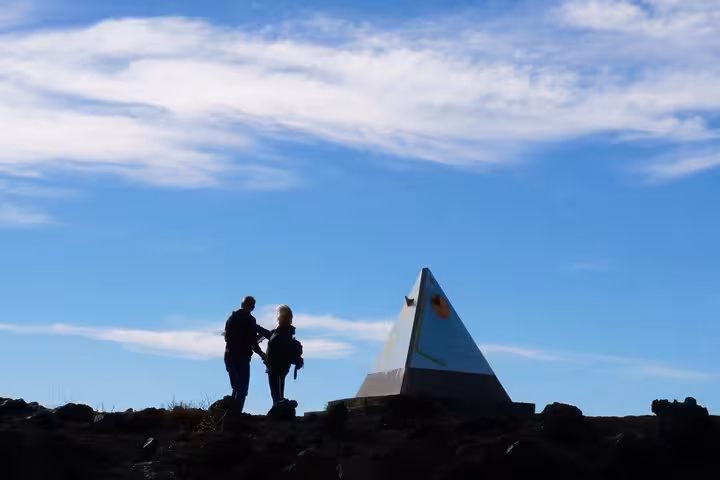Two hikers standing beside a pyramid marker on Mount Etna, enjoying panoramic views under a vibrant blue sky.