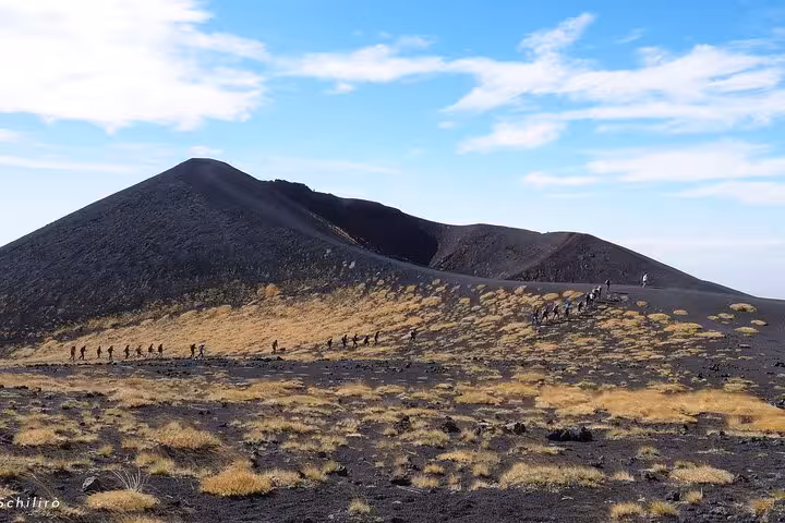 A group of hikers traverses the rugged, ash-covered landscape of Mount Etna against a backdrop of clear blue skies.