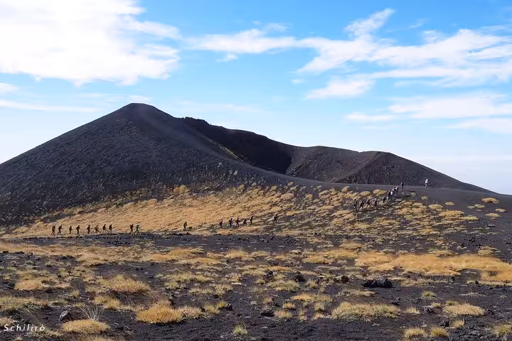 Hikers traversing the rugged volcanic landscape of Mount Etna, with clear blue skies and striking volcanic cones in view.