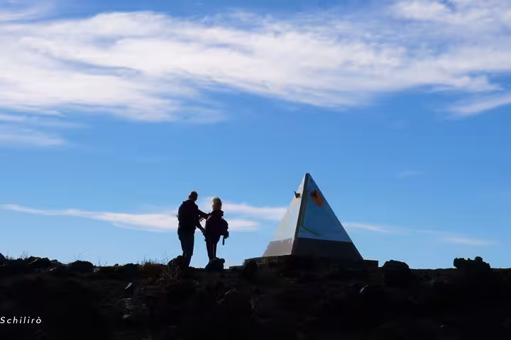 Silhouetted hikers admire a pyramid-shaped marker under a bright blue sky on Mount Etna, highlighting a scenic morning tour.