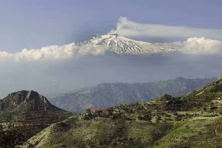 Panoramic view of Mount Etna with a plume of smoke, surrounded by lush green hills and scattered clouds.
