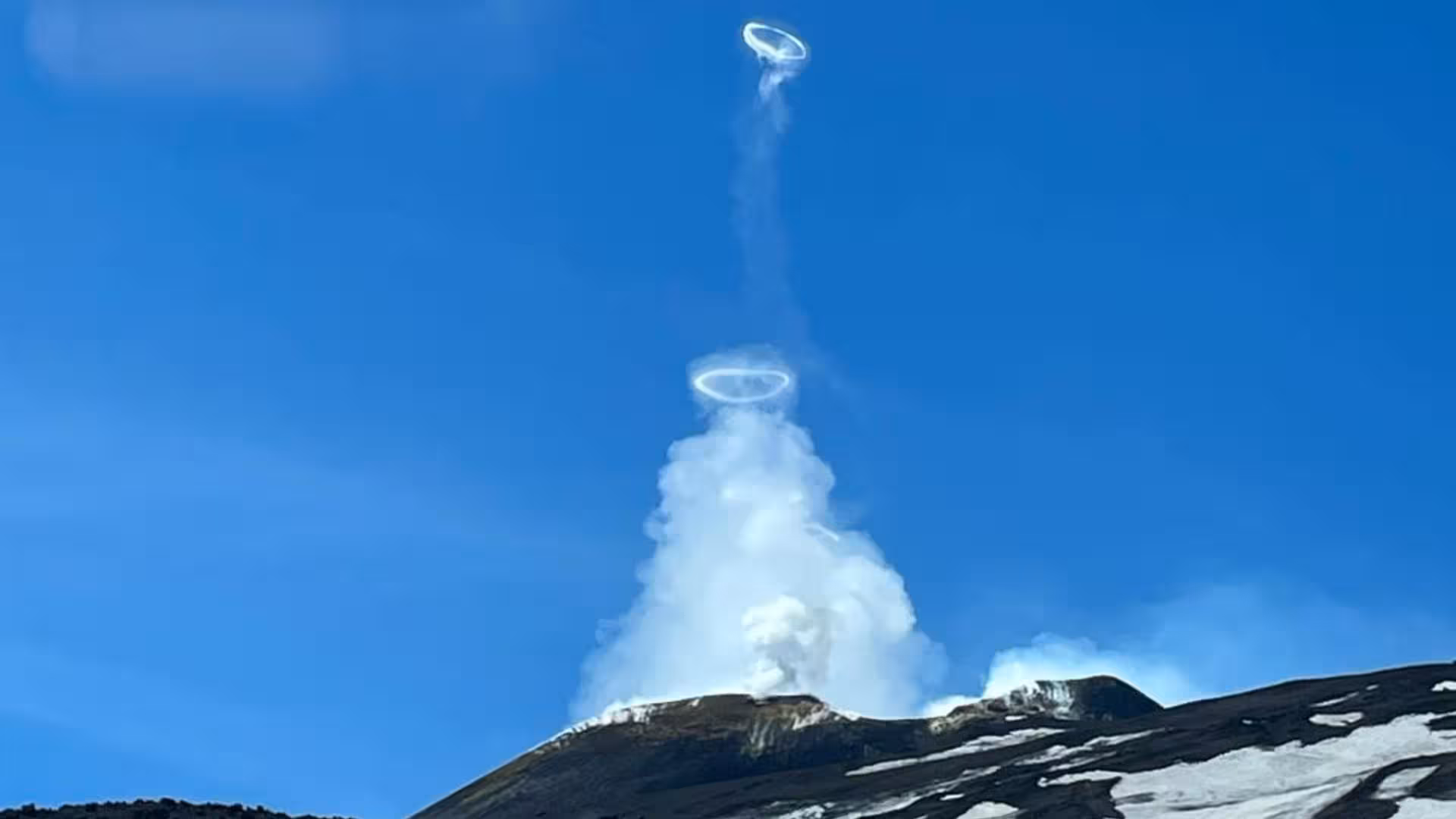 Smoke rings rise from Mount Etna's active crater against a vibrant blue sky, showcasing volcanic activity in Sicily.