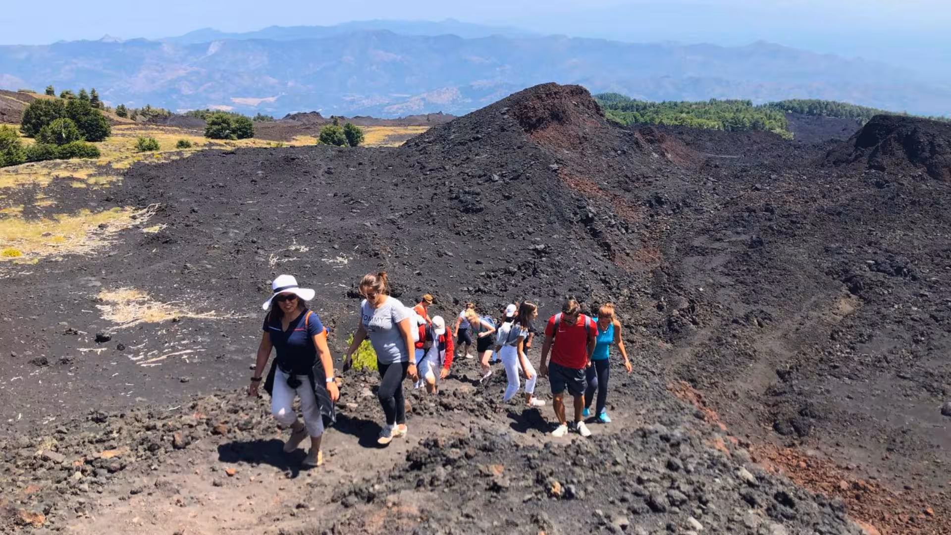 Group of hikers trekking over volcanic terrain on Mount Etna with scenic views of distant craters and landscape.