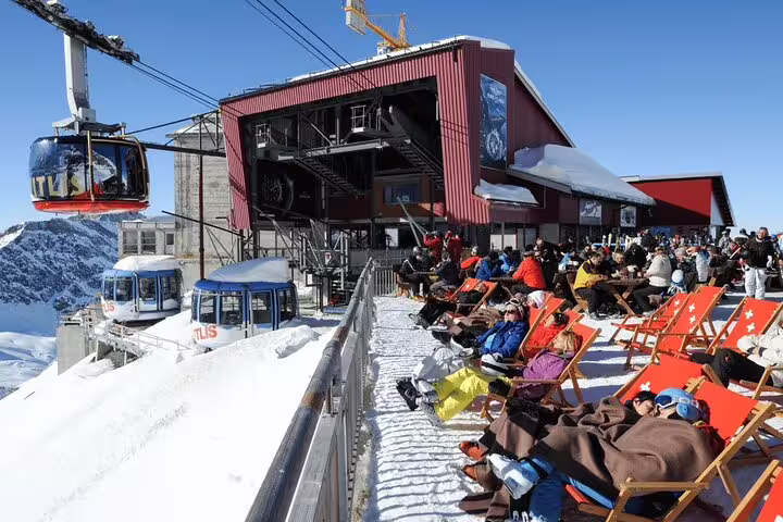 Travelers relax in deck chairs at Mount Titlis summit station, enjoying breathtaking alpine views and cable cars.