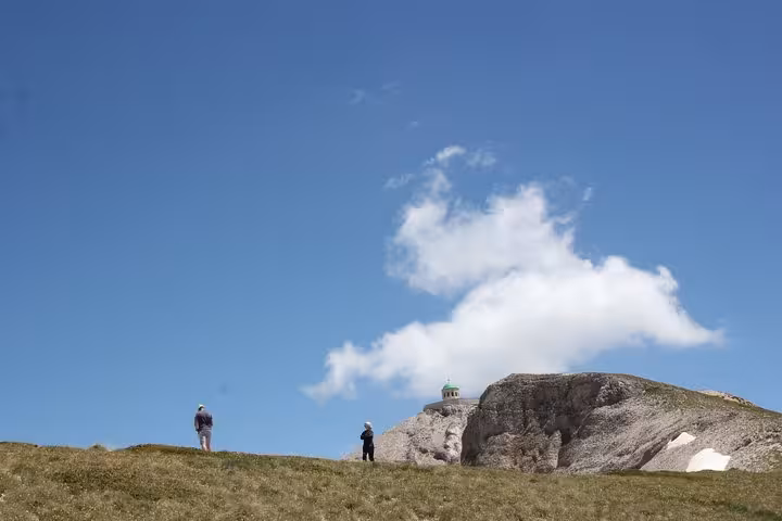 Two hikers explore grassy slopes near a rocky outcrop on Mount Tomorr under a bright blue sky, ideal for guided trips.