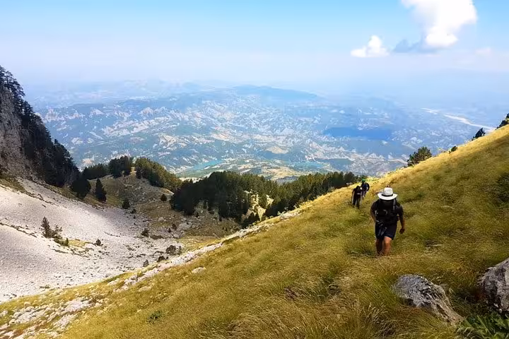 Adventurers hike uphill on lush green slopes of Mount Tomorr, overlooking expansive panoramic views of the valley below.