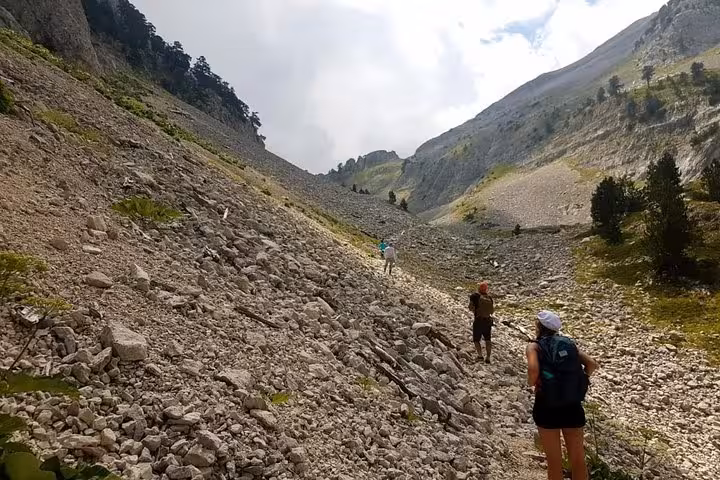 Hikers traverse a rocky path on Mount Tomorr, surrounded by rugged landscapes and distant peaks under a cloudy sky.