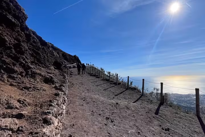 Visitors hike along the rocky pathway of Mount Vesuvius, with panoramic views of the coastline under the bright sun.