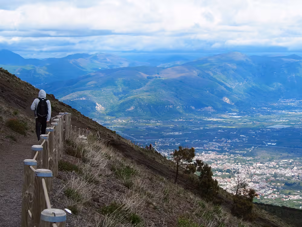 Hiker on Mt Vesuvius trail with panoramic views of lush hillsides and the town below, perfect for a Naples day trip.
