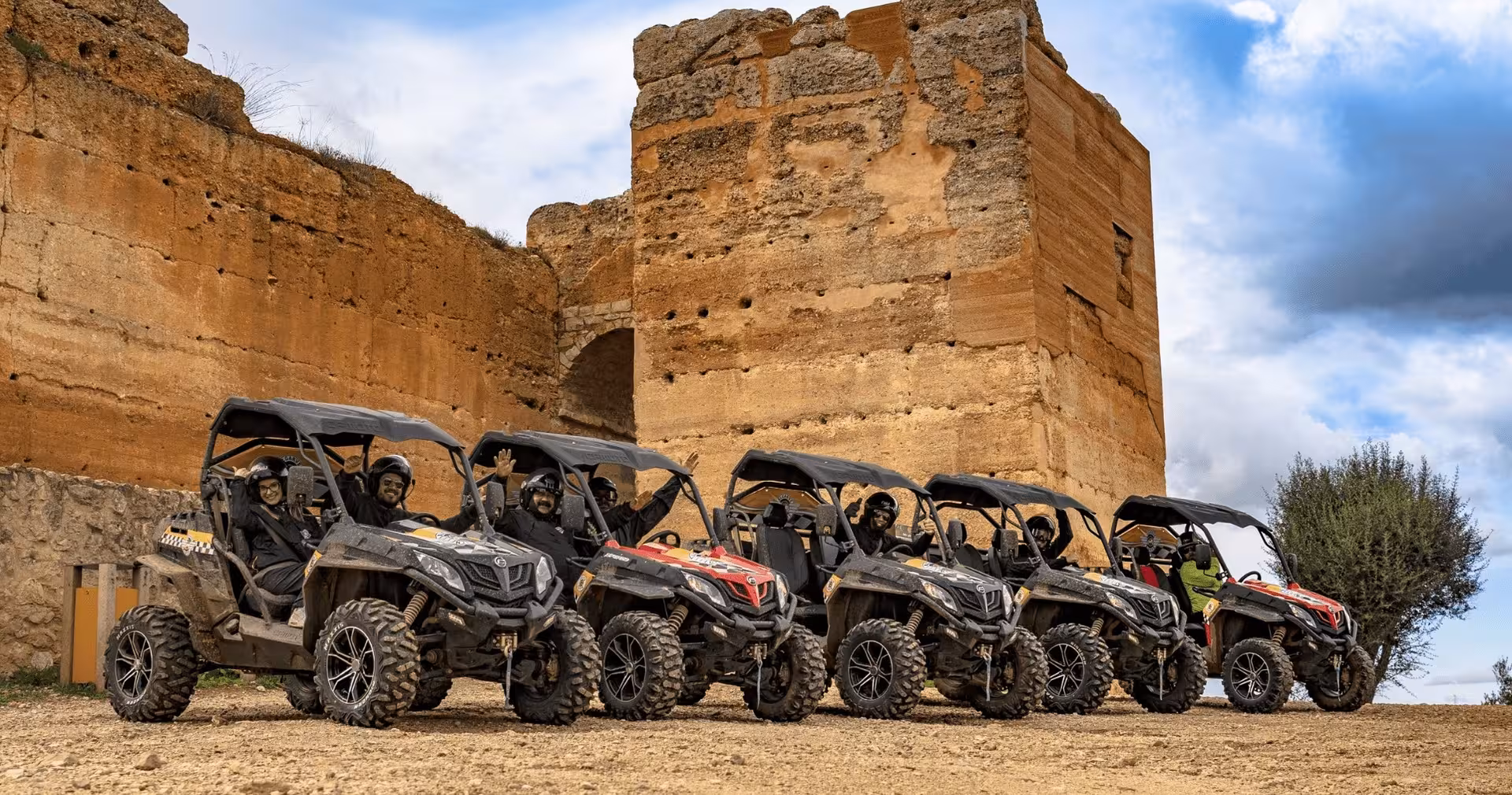 Line of muddy buggies parked by ancient stone ruins on a half-day off-road tour, with helmeted drivers waving at the camera.