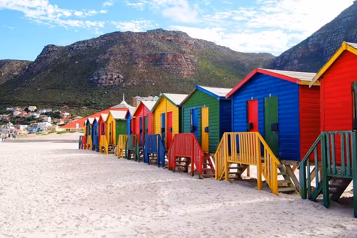 Brightly colored beach huts line the sandy shores with a mountainous backdrop at Muizenberg, Cape Town.