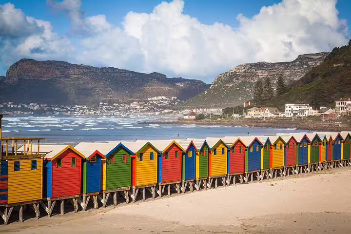 Colorful beach huts line the sandy shore with mountain backdrop at Muizenberg Beach on a Cape Town tour.