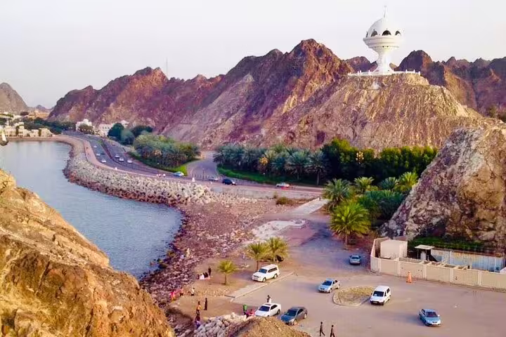 Aerial view of Muscat's coastal road with rocky mountains and iconic incense burner monument during a city tour.