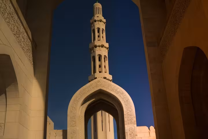 Illuminated minaret and ornate archway of Sultan Qaboos Grand Mosque at dusk on a Muscat evening taxi sightseeing tour