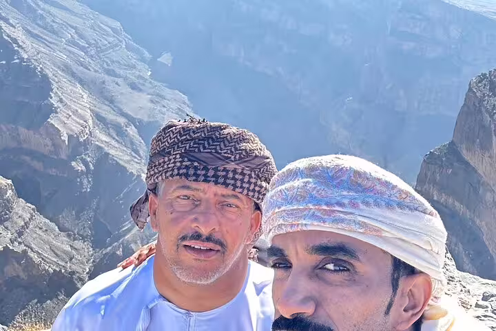 Two men in traditional attire enjoying the breathtaking views of Jabal Al Akhdar's rugged cliffs on a sunny day.