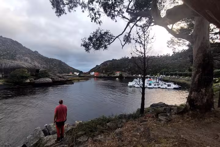 Man standing by a tranquil coastal inlet in Muxia, with boats moored and scenic hills in the background.