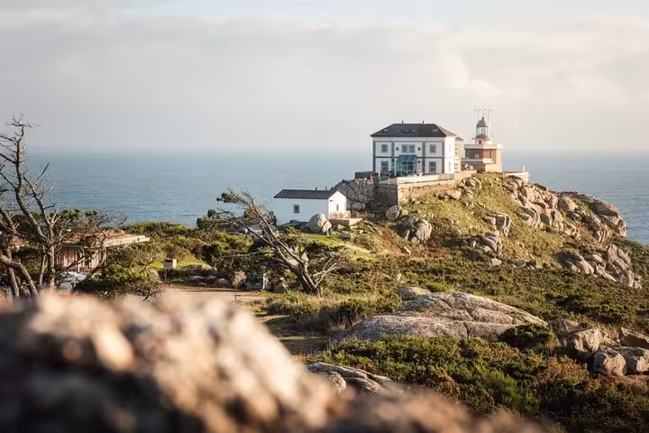 Charming coastal view of Muxia lighthouse and building atop a rocky hill with expansive sea backdrop at sunset.