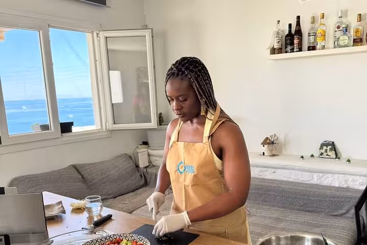 Guest chops herbs beside sea-view window during Mykonian cooking class for cruise travelers, learning authentic Greek recipes