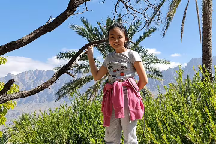 Young tourist poses amidst lush greenery and palm trees with mountain backdrop in Wakan Village on Nakhal tour.