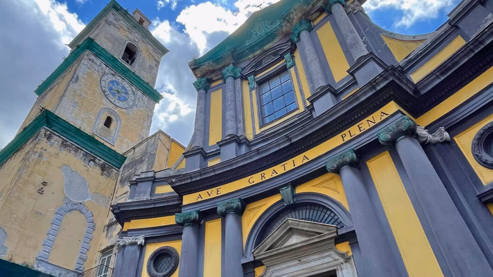 Stunning view of a historic yellow and gray church in Naples with a clock tower against a cloudy sky.