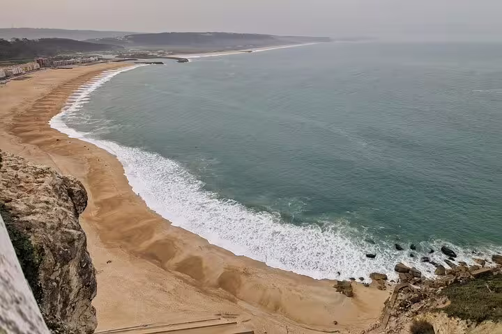 Panoramic view of Nazaré's sandy beach with rolling waves and scenic cliffs under a serene sky.