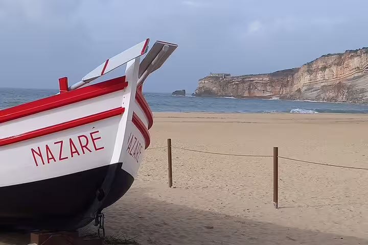 A traditional boat with 'Nazare' written on it rests on a sandy beach with cliffs and ocean waves in the background.