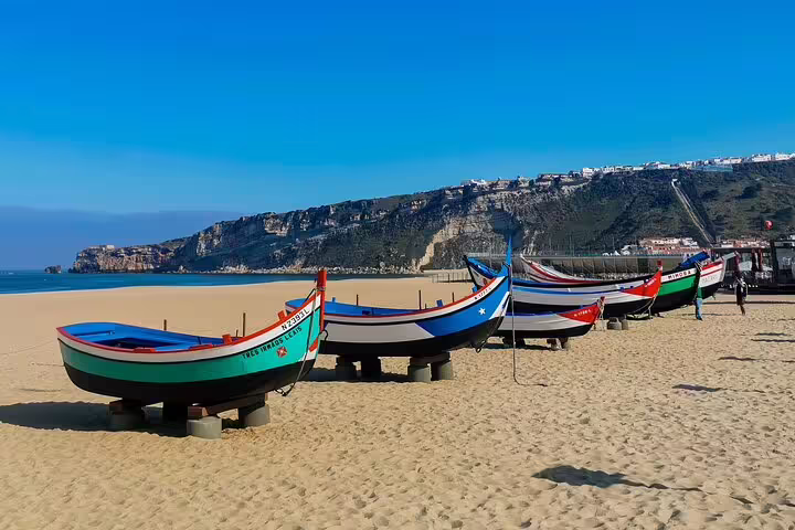 Colorful traditional fishing boats on the sandy beach of Nazaré, Portugal, with cliffs and blue sky in the background.