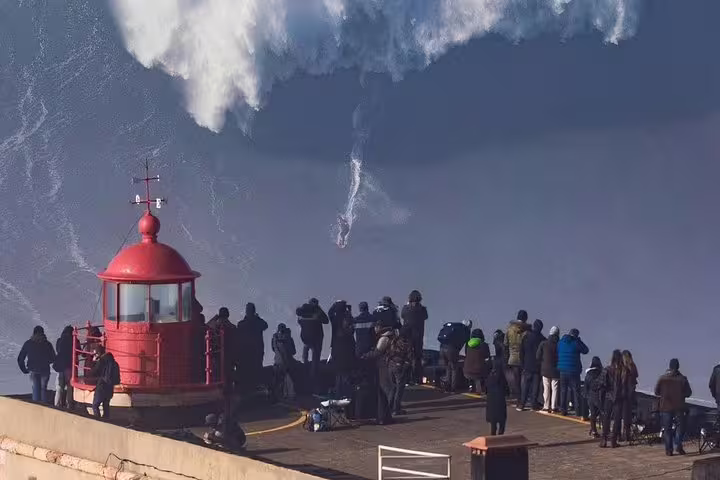 Tourists watching a surfer ride massive waves near the iconic red lighthouse in Nazaré, Portugal on a private tour of Fatima, Nazaré, and Óbidos.