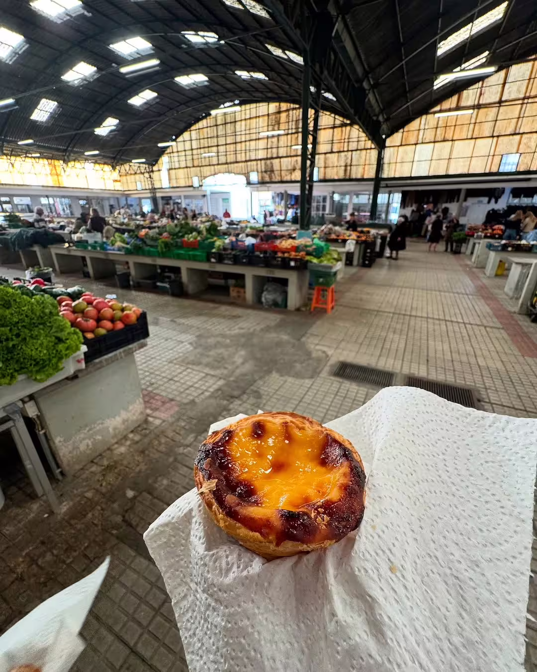 A delicious pastel de nata is held in the foreground of a vibrant market in Nazaré, capturing local culture and flavors.