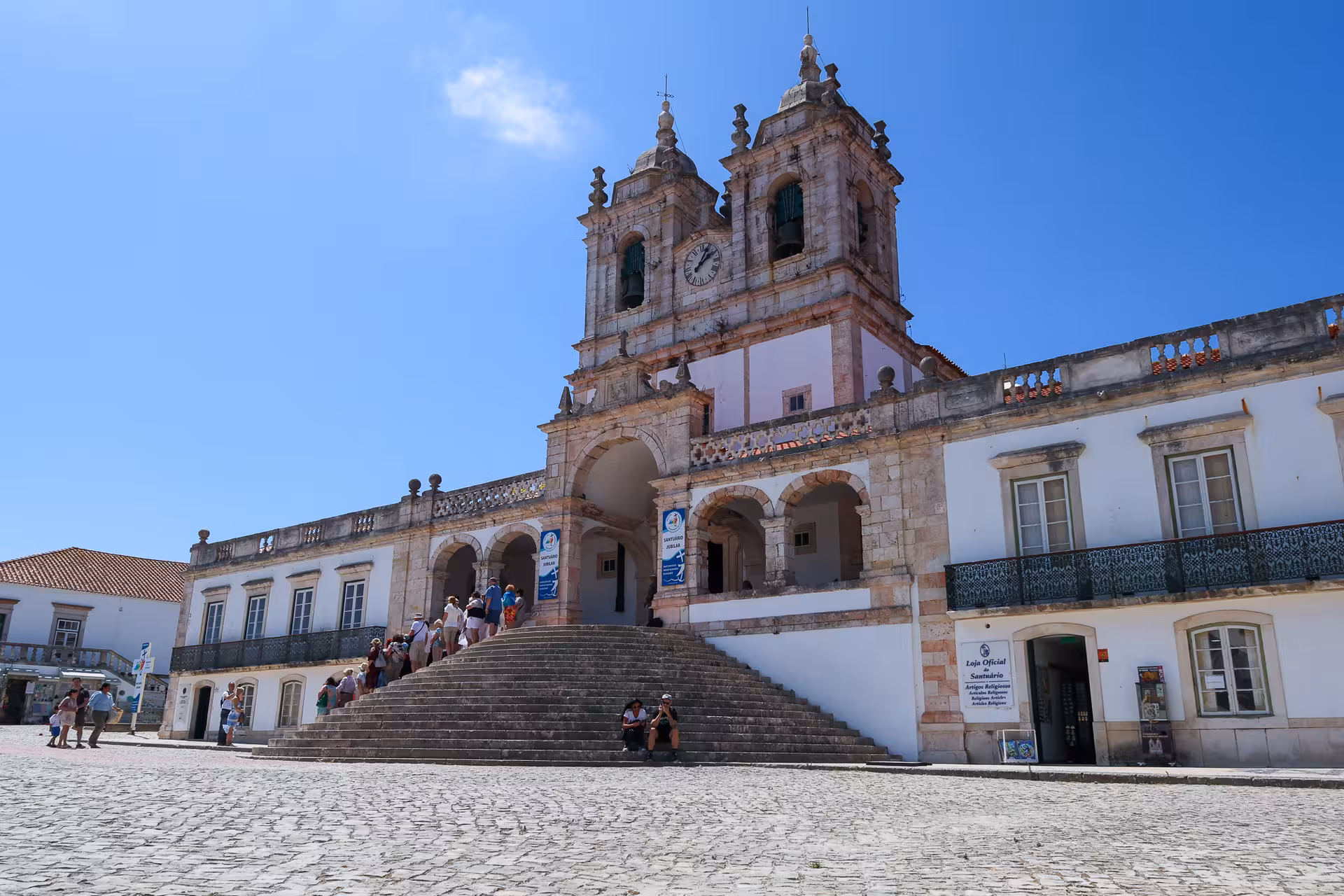 Tourists visit the historic Sanctuary of Our Lady of Nazaré under a clear blue sky in Portugal.