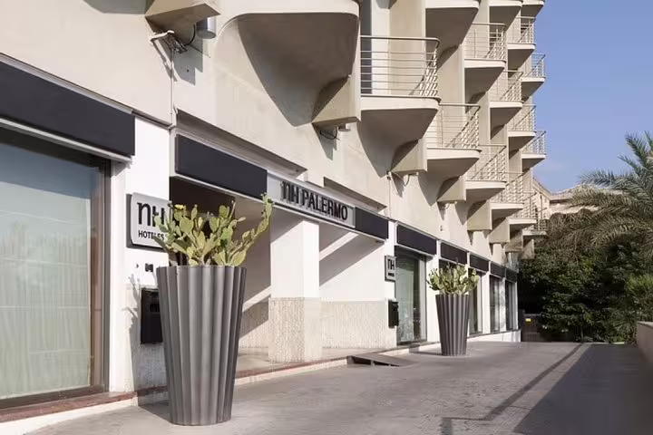 Exterior entrance of NH Palermo Hotel with modern balconies and tall planters for private airport transfer drop-off