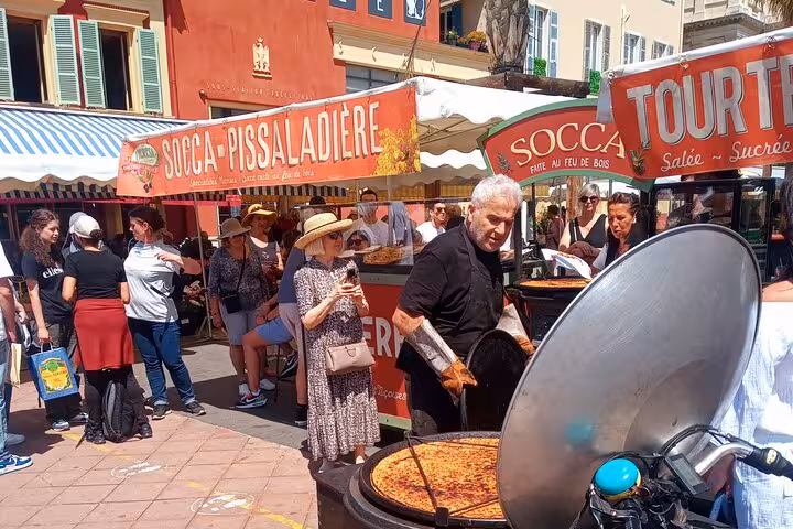 Bustling Nice market with locals enjoying traditional socca and pissaladière under vibrant awnings.