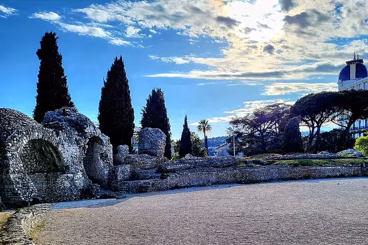 Scenic view of ancient Roman ruins in Nice with towering cypress trees and a bright blue sky.