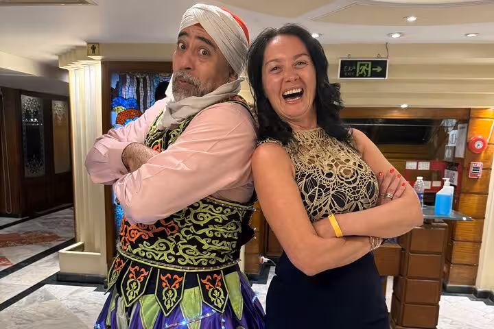 Smiling woman and performer in colorful attire pose on a Nile cruise, highlighting cultural entertainment.