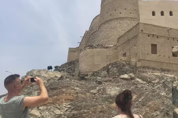 Tourists capture the majestic stone architecture of Nizwa Fort, Oman, showcasing its historical significance on a rocky hill.