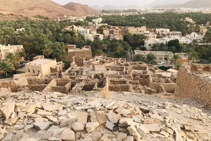 Panoramic view of Nizwa's old town ruins surrounded by lush palm groves and modern buildings in the distance.