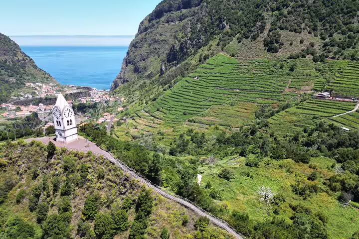 Aerial view of lush terraced hills and a charming chapel overlooking the sea in Northeast Madeira, ideal for scenic guided tours.