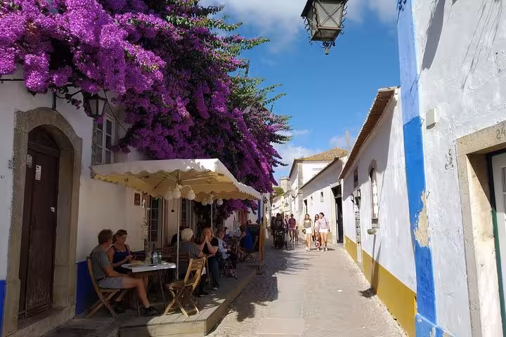 People enjoying a sunny café under purple bougainvillea on a quaint Óbidos street, ideal for a private Portugal tour.