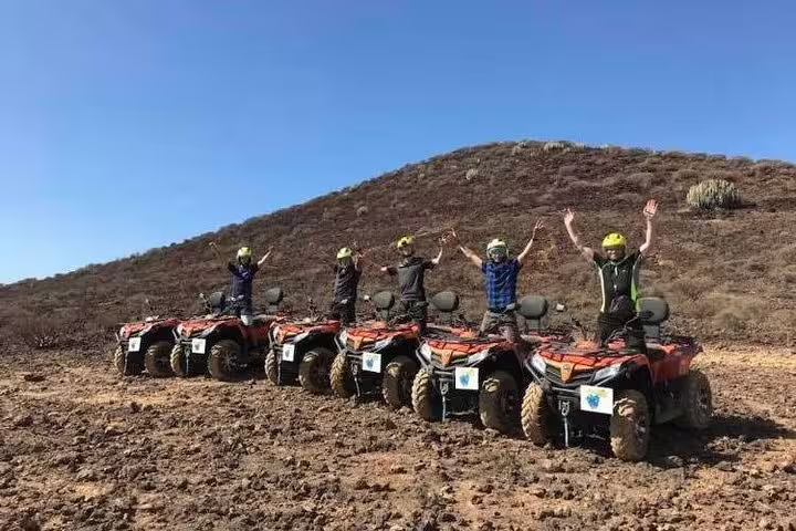 Enthusiastic group celebrates atop quad bikes against a backdrop of rocky hills and clear blue sky.