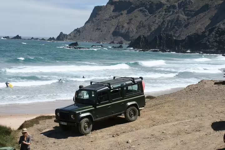 Off-road vehicle overlooking stunning Algarve beach with surfers enjoying the waves, perfect for adventure tours.
