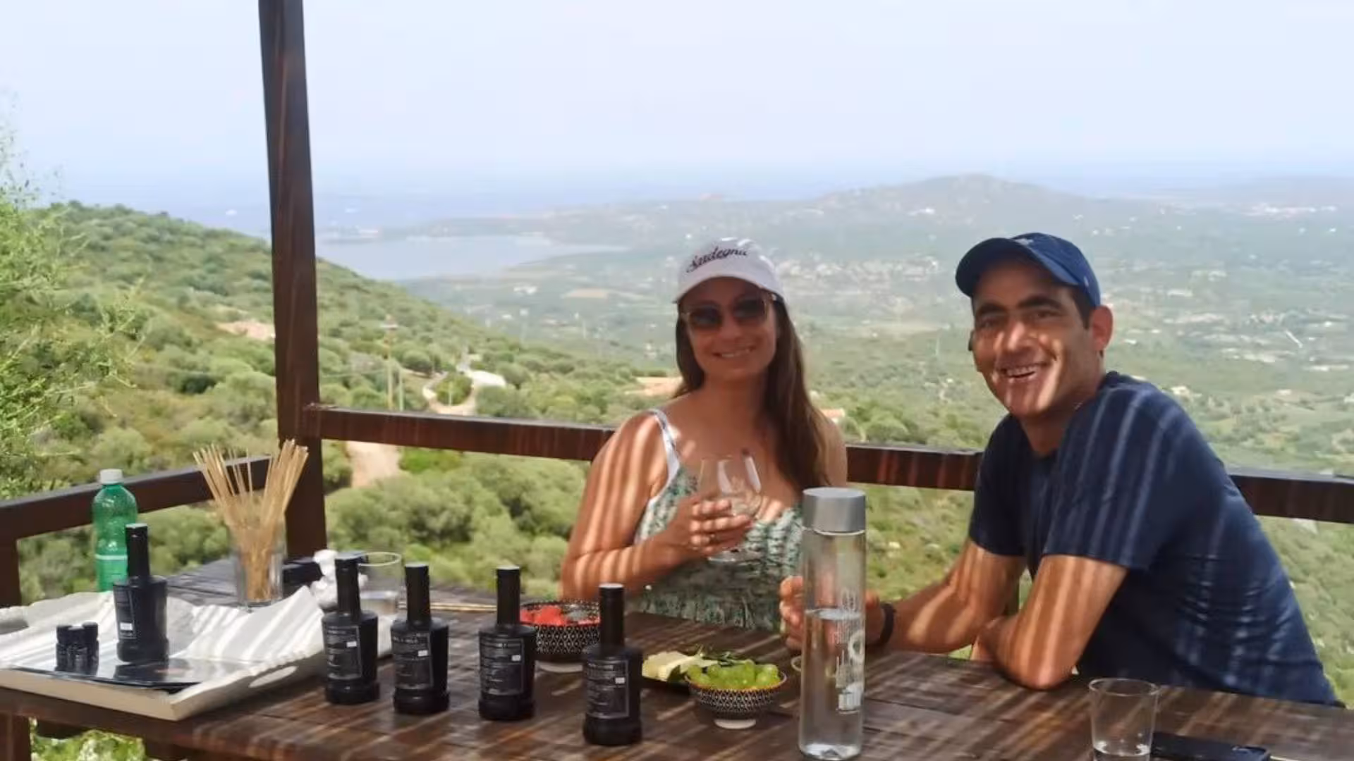 Visitors enjoying myrtle liqueur tasting with stunning views of Olbia's hillside and sea backdrop.